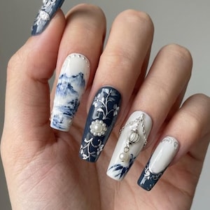 May include: A hand displaying long, square-shaped artificial nails. The nails feature intricate designs, including a landscape scene, floral patterns, and decorative elements like pearls and chains. The color palette includes shades of blue, white, and silver.