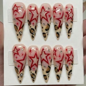 May include: Set of ten long, almond-shaped false nails. Each nail features a design with a red star, small rhinestones, and a leopard print pattern at the tip. The nails are displayed on a white card.