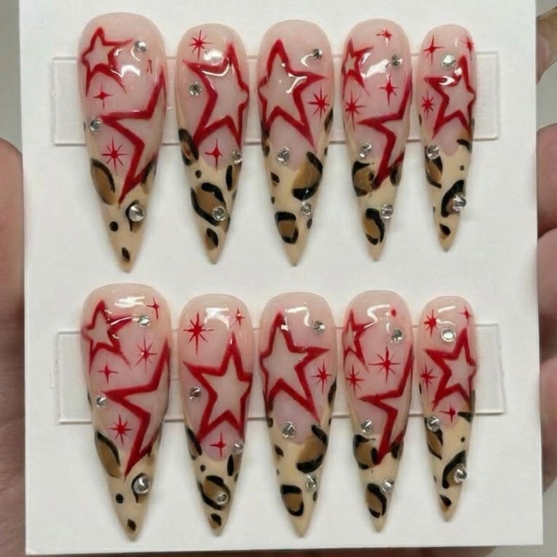 May include: Set of ten long, almond-shaped false nails. Each nail features a design with a red star, small rhinestones, and a leopard print pattern at the tip. The nails are displayed on a white card.
