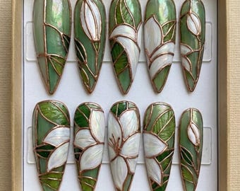 May include: A collection of ten almond-shaped press-on nails with a stained-glass effect. Each nail showcases a green and white floral design, outlined in a copper hue. The nails are presented in a rectangular box.