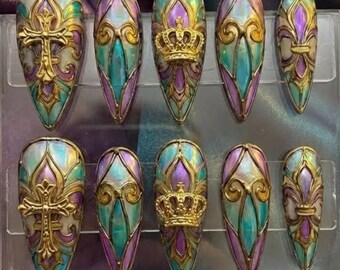 Può includere: Un set di dieci unghie finte a forma di mandorla. Ogni unghia presenta un design con dettagli dorati, tonalità iridescenti di verde acqua, viola e bianco. Alcune unghie hanno un design a croce, corona o giglio.