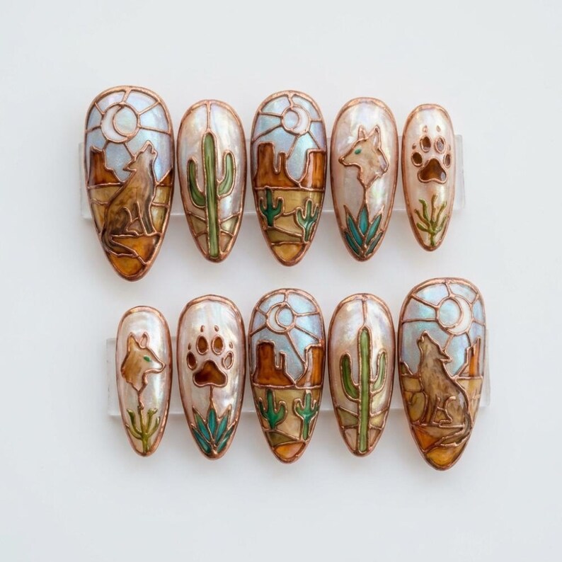 Puede incluir: Un conjunto de diez u&ntilde;as postizas ovaladas con dise&ntilde;os tem&aacute;ticos del desierto. Cada u&ntilde;a presenta una base nacarada con im&aacute;genes de cactus, lobos aullando, huellas de patas y paisajes des&eacute;rticos en tonos marrones, verdes y azules.
