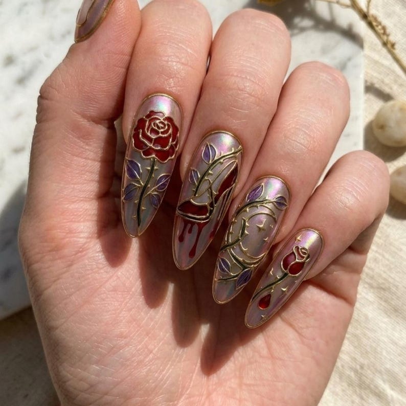 May include: Long, oval-shaped nail art with a pearlescent finish. Each nail features a different design, including a red rose, a high-heeled shoe, and rose vines, all outlined in gold. The designs are painted on a light pink base.