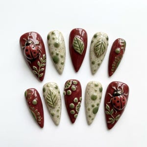 May include: A set of ten press-on nails featuring a glossy finish in deep red and cream colors. The designs include raised 3D ladybugs, green leaf motifs, and small green polka dots arranged in various patterns across the almond-shaped nail tips.