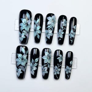 May include: A set of ten long, coffin-shaped press-on nails with a glossy black base. Each nail features hand-painted light blue floral designs with iridescent highlights and small, clear rhinestones centered on some of the flower petals.