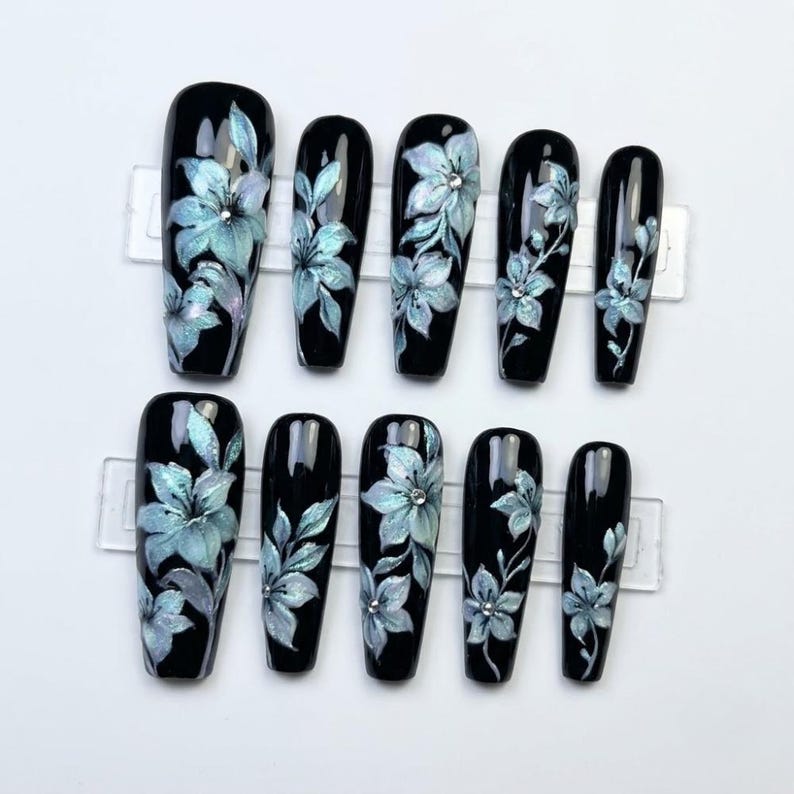 May include: A set of ten long, coffin-shaped press-on nails with a glossy black base. Each nail features hand-painted light blue floral designs with iridescent highlights and small, clear rhinestones centered on some of the flower petals.