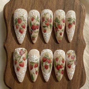 May include: Set of ten almond-shaped false nails. Each nail features a hand-painted design of strawberries with green leaves and white decorative swirls. The nails have a beige base color and are arranged on a wooden surface.