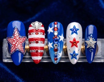 May include: A set of five almond-shaped press-on nails featuring patriotic designs. The nails display red, white, and blue colour schemes with embellishments including a leopard print star, horizontal stripes, rhinestones, and raised star accents.