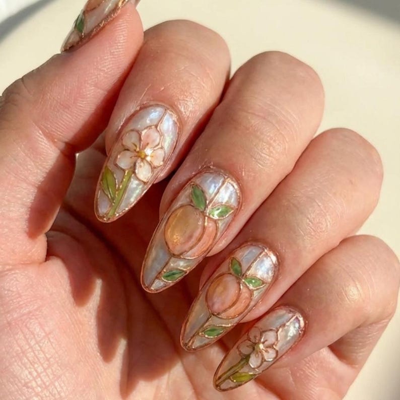 K&ouml;nnte beinhalten: Mandelf&ouml;rmige Nagelkunst mit perlmuttfarbener Basis, Pfirsich- und Blumendesigns. Die N&auml;gel haben eine goldene Kontur und sind mit pfirsichfarbenen Fr&uuml;chten und wei&szlig;en Blumen mit gr&uuml;nen Bl&auml;ttern bemalt.