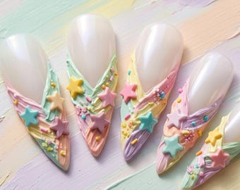 May include: Five almond-shaped nail art tips featuring a V-shaped French manicure design. The tips are decorated with textured pastel paint strokes in mint, yellow, pink, and lavender, accented with small colourful star charms and tiny sprinkles.