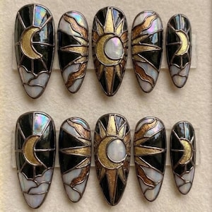 May include: A set of ten press-on nails with a celestial design. Each nail features a black background with gold, white, and iridescent accents. The designs include sun and moon motifs, resembling stained glass.