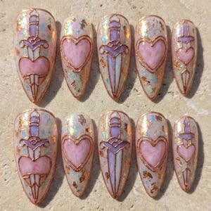 May include: Set of ten almond-shaped false nails. Each nail features a design with a pink heart or a sword piercing a heart, outlined in copper-colored metal. The nails have a pearlescent base with gold foil accents.
