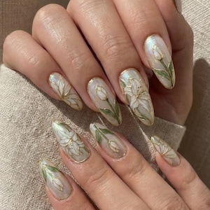 May include: A hand with long, almond-shaped nails. Each nail is painted with a pearlescent base and features a delicate floral design in white and green, outlined with gold. The designs include lilies and tulips.