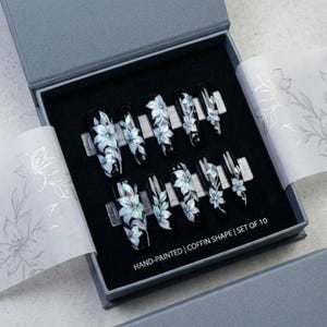 May include: A set of ten coffin-shaped press-on nails in a gray box. Each black nail features hand-painted light blue and white floral designs. The box interior includes text reading HAND-PAINTED | COFFIN SHAPE | SET OF 10, with floral tissue paper.