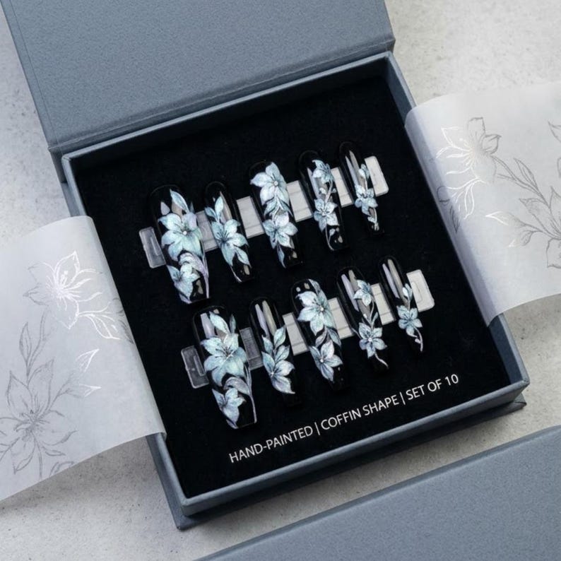 May include: A set of ten coffin-shaped press-on nails in a gray box. Each black nail features hand-painted light blue and white floral designs. The box interior includes text reading HAND-PAINTED | COFFIN SHAPE | SET OF 10, with floral tissue paper.