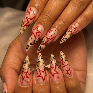 May include: Long, pointed fingernails with a clear base, adorned with red star designs and small rhinestones. The tips feature a leopard print pattern, creating a bold and stylish nail art design.
