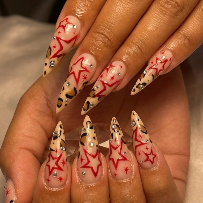 May include: Long, pointed fingernails with a clear base, adorned with red star designs and small rhinestones. The tips feature a leopard print pattern, creating a bold and stylish nail art design.
