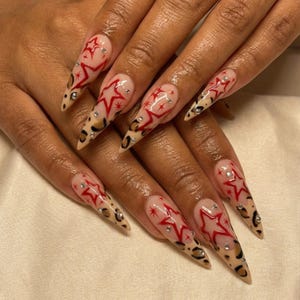 May include: Long, pointed fingernails with a clear base, red star designs, and leopard print tips. Each nail features a red star outline with small, sparkling embellishments. The nails are a fashionable accessory.