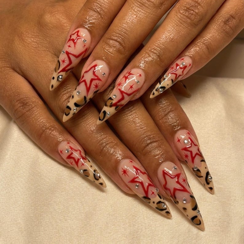 May include: Long, pointed fingernails with a clear base, red star designs, and leopard print tips. Each nail features a red star outline with small, sparkling embellishments. The nails are a fashionable accessory.