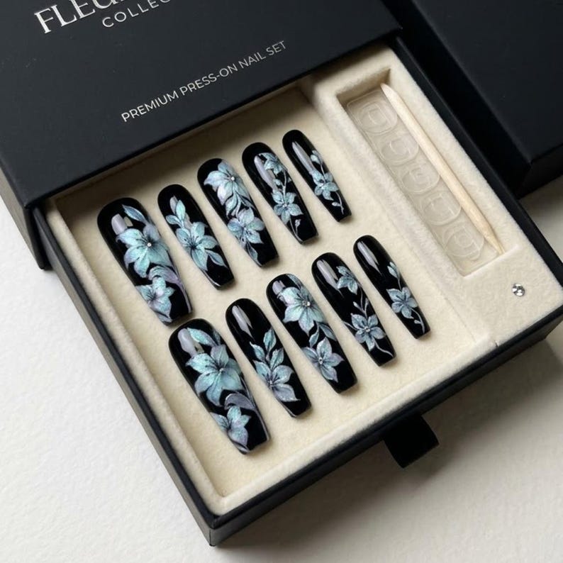 May include: A set of ten black press-on nails with a glossy finish, each featuring hand-painted light blue floral designs with small silver accents. The nails are arranged in a black box labeled PREMIUM PRESS-ON NAIL SET with a wooden cuticle stick.