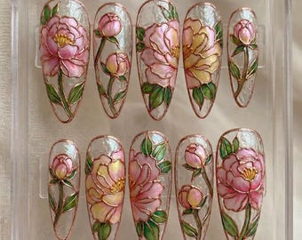 May include: A set of ten artificial nails with a floral design. Each nail features a pink and yellow peony flower with green leaves, outlined in gold. The nails are displayed in a clear plastic case.