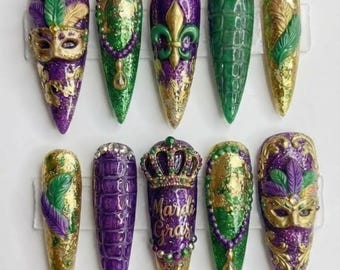 May include: A set of ten long, pointed artificial nails with a Mardi Gras theme. The nails feature glitter, masks, feathers, crowns, and the words "Mardi Gras". Colors include purple, green, gold, and silver.