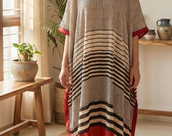 May include: A woman wearing a beige, black, and red striped caftan dress. The dress has a V-neck and short sleeves with red trim. The hemline and sleeve edges are trimmed in red. The dress is made of a lightweight fabric.