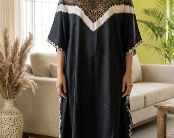 May include: A black caftan dress with a V-neck and short sleeves. The top of the dress features a brown and white patterned design. The dress has white trim along the sleeves and hemline. The woman is standing in a well-lit room.