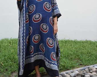 May include: A navy blue kaftan dress featuring a repeating circular design in red, white, and black. The dress has a V-neck and wide sleeves. The model is wearing sunglasses and earrings. The dress is worn with black trousers and red embroidered shoes.