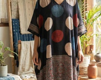 May include: A black kaftan dress with a V-neck and large, colourful polka dots. The dress has a patterned border at the hem and sleeves. The model is standing in a well-lit room with natural light.