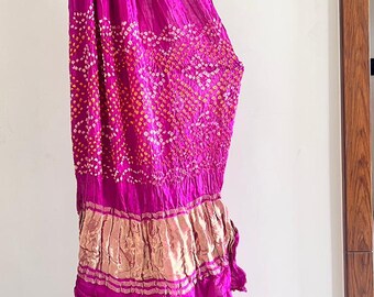 May include: A vibrant magenta fabric featuring a traditional tie-dye pattern with small white and yellow dots. The textile includes a wide, horizontal band of metallic gold fabric with a pleated texture, positioned towards the lower section of the drape.