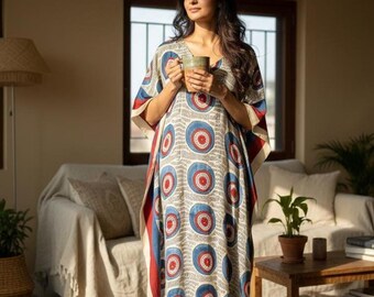 May include: A long kaftan dress featuring a pattern of red and blue circles on a cream base. The dress has a V-neck and wide sleeves. The person is holding a mug. The dress is suitable for relaxed wear. The dress is approximately 150 cm in length.