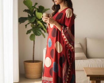 May include: A red kaftan dress with large polka dots in various colours. The dress has a V-neck and patterned trim. The person is holding a mug. The dress is suitable for casual wear.