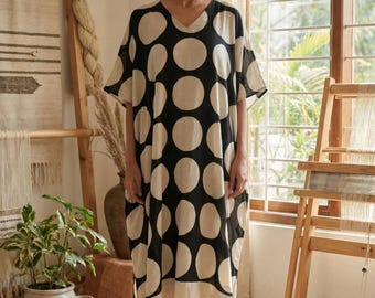 May include: A black and cream polka-dot dress with a V-neck and short sleeves. The dress has a loose fit. The model is wearing sandals. The background includes a window, wooden furniture, and decorative items, creating a relaxed atmosphere.