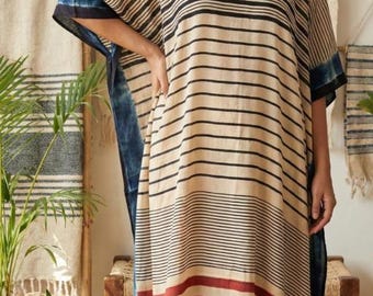 May include: A beige, striped caftan dress with black and red horizontal stripes. The dress has a V-neck and wide sleeves with blue accents. The model is standing in front of a neutral background.