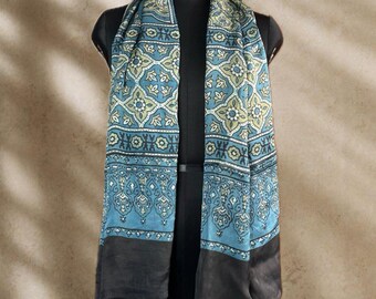 May include: A rectangular scarf featuring a teal and sage green geometric floral pattern on the upper section, transitioning into a solid dark charcoal grey border at the bottom. The fabric displays a smooth texture with a subtle sheen throughout.