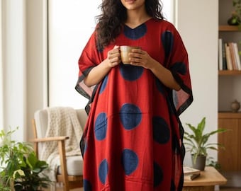 May include: A red kaftan dress with large navy blue polka dots. The dress has a V-neck and black trim. The person is holding a mug. The dress is a comfortable, loose-fitting garment, suitable for lounging or casual wear.