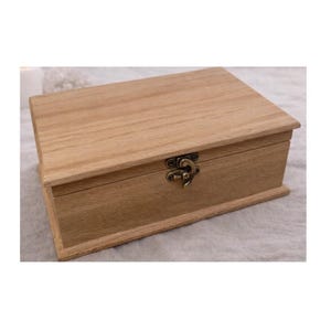 Puede incluir: Una caja rectangular de madera con tapa con bisagras y cierre metálico. La caja es de madera clara con veta visible. El cierre es de metal oscuro de estilo antiguo. La caja está cerrada y se encuentra sobre una superficie de color neutro.