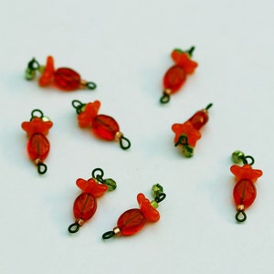 May include: Close-up of several handmade orange and red flower-shaped beads with green accents. Each bead has a small loop for stringing. The beads are scattered on a white surface, ideal for jewelry making or craft projects.