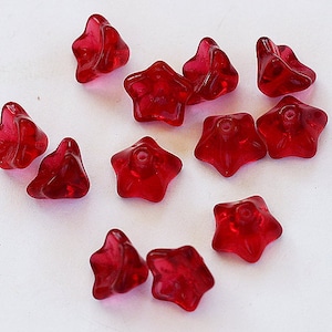 May include: A collection of translucent, star-shaped beads in a vibrant red hue. Each bead has a five-point design with a small hole, suggesting they are for stringing. The beads are scattered on a white surface, showcasing their glossy texture.