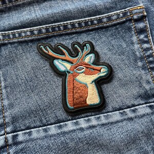 May include: Embroidered patch featuring a deer head with antlers. The patch has a black border and is sewn onto a blue denim pocket. The deer is brown, beige, and light blue.