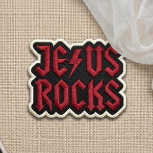 May include: Embroidered patch with the words "JESUS ROCKS" in red, outlined in black and white. The patch features a lightning bolt in place of the letter "I". The patch is on a tan fabric background.