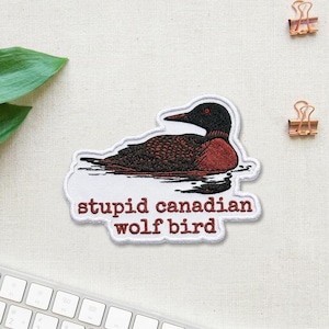 May include: Embroidered patch featuring a stylized bird with a black head, red neck, and brown body, floating on water. The patch includes the text "stupid canadian wolf bird" in a red font, outlined in white.