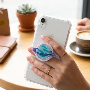 May include: A white smartphone with a clear case and a planet-themed phone grip. The grip features a blue and green planet design with a purple ring. The phone is held near a cup of coffee and a small potted plant.