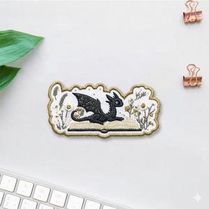 May include: Embroidered patch featuring a black dragon with gold accents, sitting on an open book. The patch is surrounded by embroidered flowers and foliage, with a gold border. The design is on a white background.