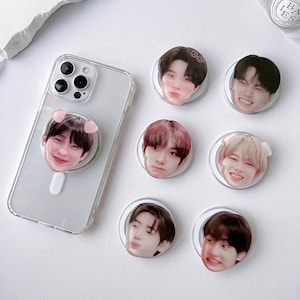 May include: A clear phone case with a pop-up grip featuring a smiling face with pink pig ears. Several round pop-up grips with different smiling faces are scattered around the phone. The grips have a white border and are on a white surface.