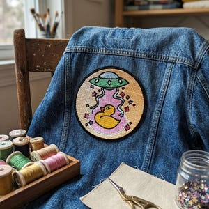 May include: A denim jacket with a large, round patch featuring a yellow rubber ducky inside a pink and gold UFO scene. The patch is embroidered. Sewing supplies, including spools of thread and scissors, are in the foreground.