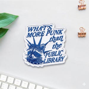 May include: Embroidered patch with a blue raccoon wearing a studded collar and a crown of spikes. The patch features the text "WHAT'S MORE PUNK than the PUBLIC LIBRARY" in blue lettering on a white background.