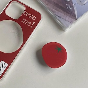 May include: A red phone case with the text "Breeze me!" and a tomato design. A red tomato-shaped phone grip with a green star design is next to the phone case. A clear CD case is in the background.