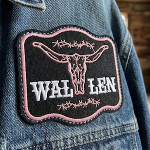 May include: Close-up of a denim jacket with a black patch featuring a pink steer skull design and the word "WAL LEN" in white. The patch has a pink border and barbed wire accents, adding a Western aesthetic.